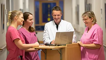 Ärztlicher Mitarbeiter und Pflegekräfte stehen im Flur an einem Stehpult und besprechen Informationen am Laptop.