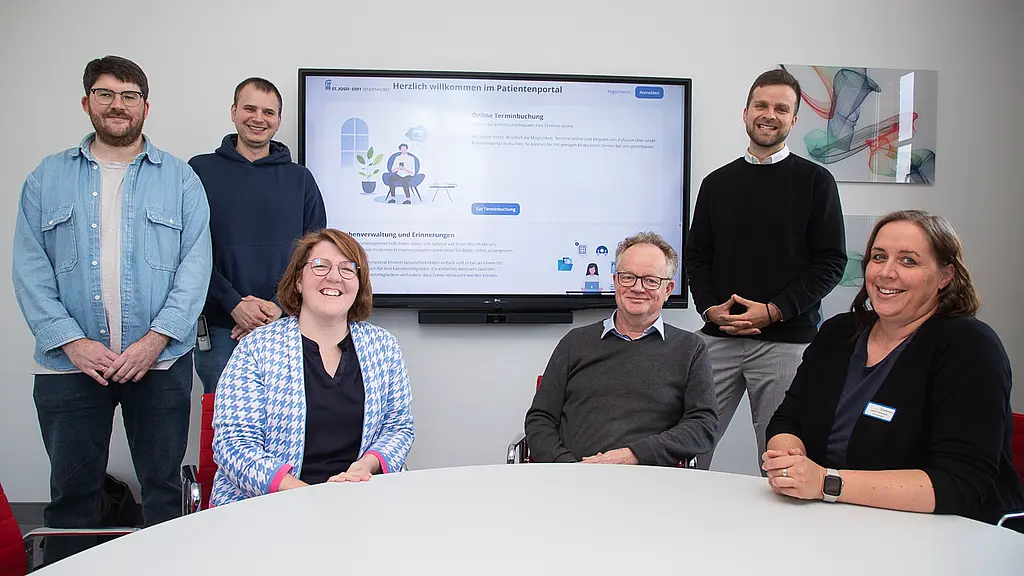 Mitarbeitende sitze am Tisch vor einem Bildschirm, auf dem das Patientenportal zu sehen ist.