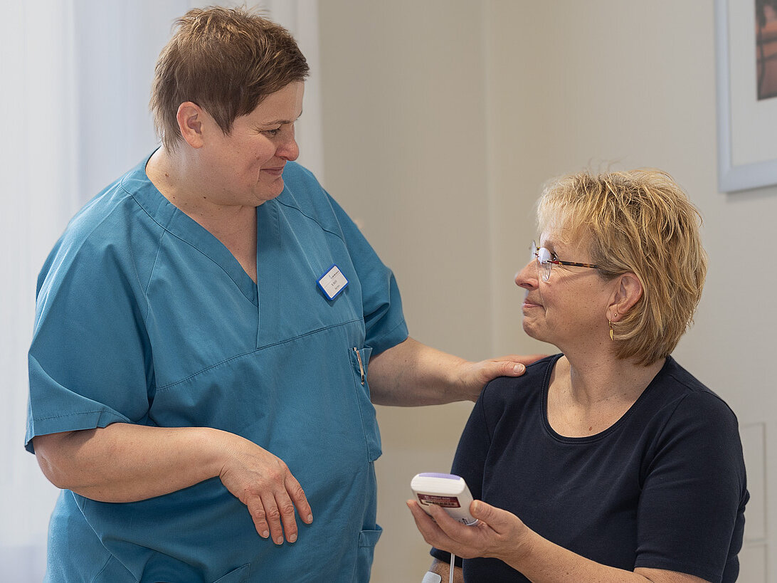 Eine Pflegefachperson erklärt einer Patientin die Anwendung eines TENS-Geräts und legt dabei beruhigend die Hand auf ihre Schulter.