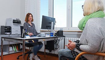 Eine Mitarbeiterin der Klinik für Ambulante Operationen & Sporttraumatologie überreicht einer Patientin am Empfang Unterlagen.