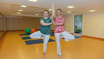 Zwei Therapeutinnen machen gemeinsam eine stehende Yoga-Übung nebeneinander.