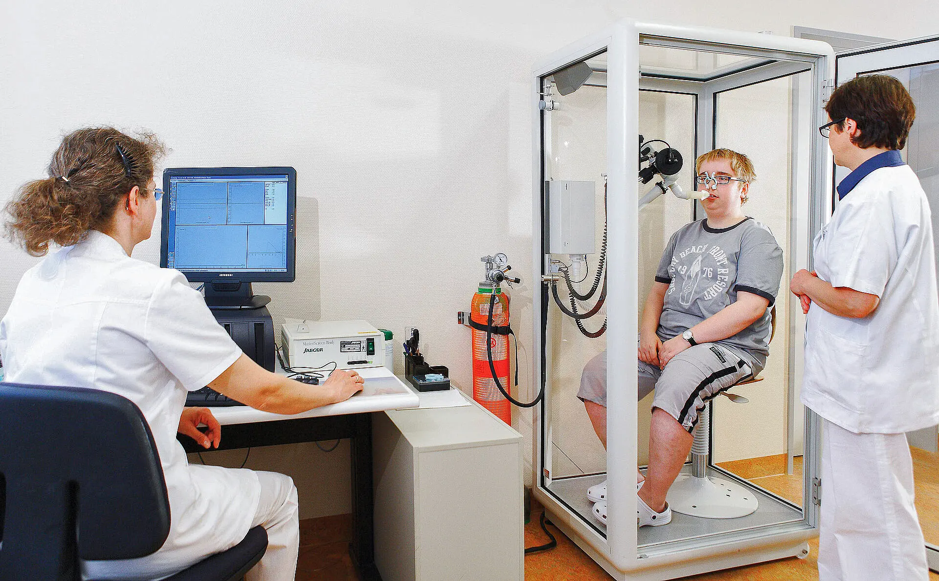 Patient sitzt in einer geschlossenen Messkabine und atmet über ein Mundstück, während zwei medizinische Fachkräfte die Untersuchung begleiten.