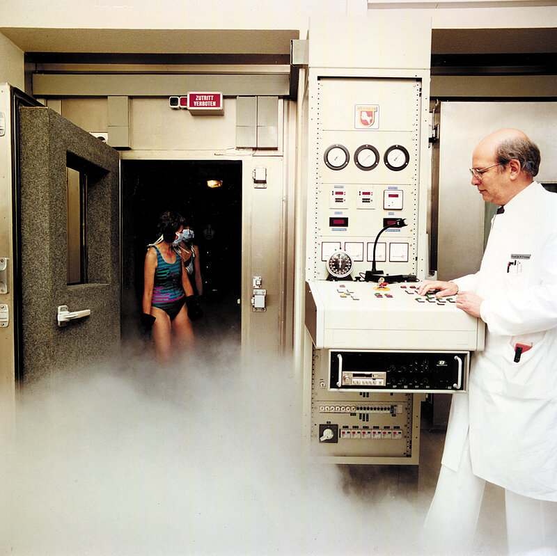 Historische Aufnahme einer medizinischen Kältekammer: Eine Patientin steht in der geöffneten Kammer, ein Arzt bedient das Steuerpult.