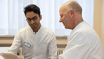Zwei Ärzte sitzen nebeneinander und betrachten gemeinsam ein Dokument. Einer der Ärzte hält das Papier in der Hand.