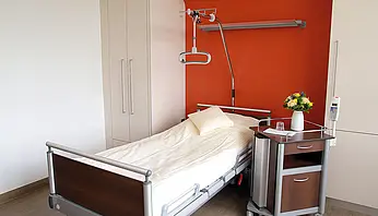 Pflegebett mit Nachttisch, Infusionsständer und Schrank vor einer farblich gestalteten Wand.