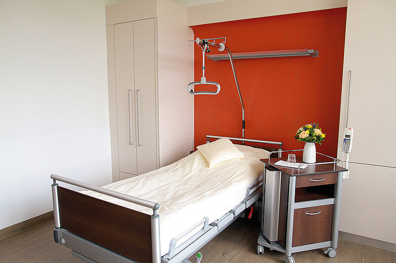 Pflegebett mit Nachttisch, Infusionsständer und Schrank vor einer farblich gestalteten Wand.