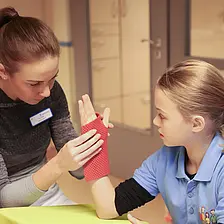 Ergotherapeutische Fachkraft überprüft die Handstellung eines Mädchens mit Kinderrheuma mithilfe einer roten Handmanschette in einem Therapieraum.