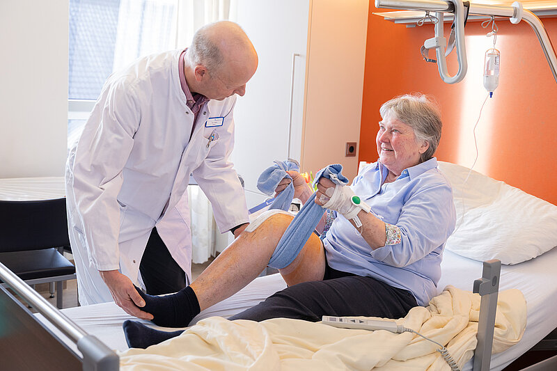 Eine Patientin sitzt im Bett und führt eine Beinübung mit einem Stofftuch durch. Ein Arzt steht neben ihr und hilft bei der Bewegung.