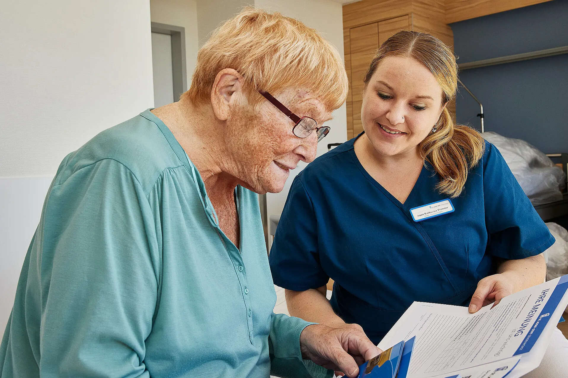 Eine Pflegefachkraft erklärt einer älteren Patientin Unterlagen und zeigt auf ein Formular, während beide aufmerksam lesen.