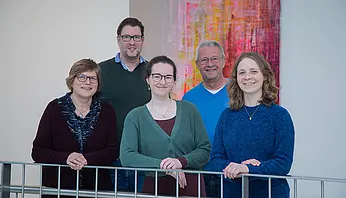 Fünf Männer und Frauen stehen lächelnd vor einem Bild und schauen in die Kamera.