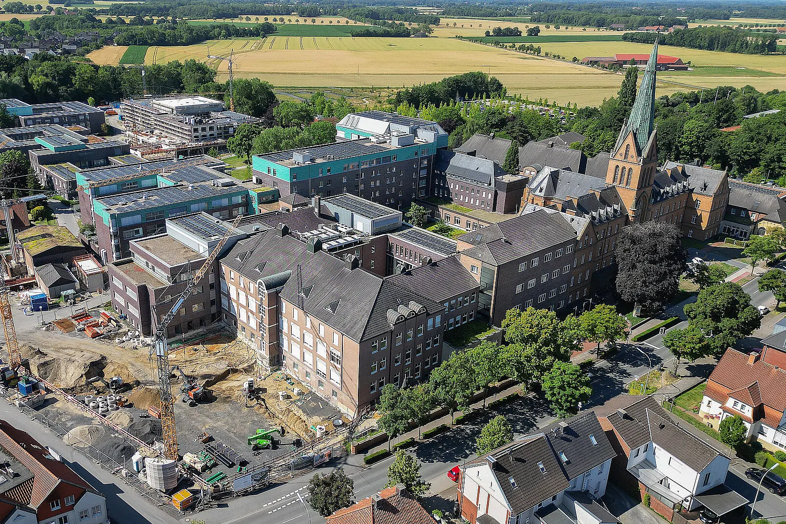 Luftaufnahme des Campus des St. Josef-Stift Sendenhorst mit mehreren Klinikgebäuden, Baustellenbereichen, einer Kirche und angrenzender Wohn- und Landschaftsfläche.