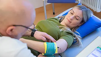 Eine Patientin liegt auf einer Therapieliege, während eine Fachkraft eine türkis-gelbe Elektrodenbandage am Unterarm anlegt. Ein Kabel führt zu einem angeschlossenen Therapiegerät.
