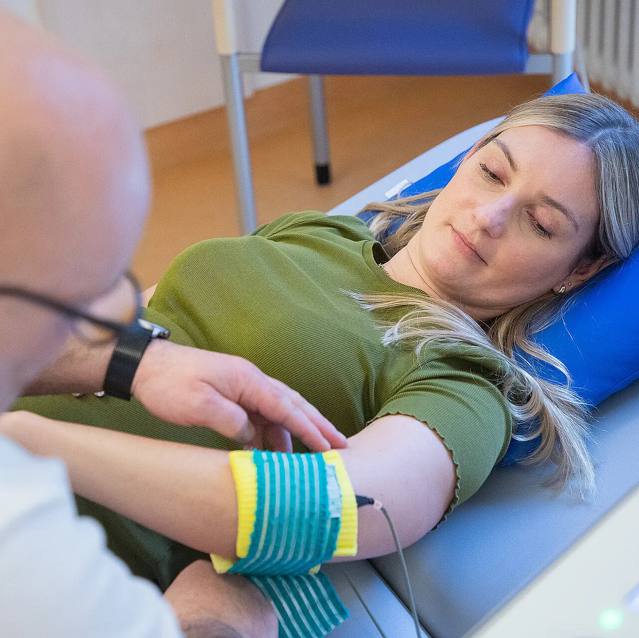 Eine Patientin liegt auf einer Therapieliege, während eine Fachkraft eine türkis-gelbe Elektrodenbandage am Unterarm anlegt. Ein Kabel führt zu einem angeschlossenen Therapiegerät.