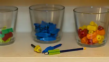 Drei Gläser mit farblich sortierten Griff- und Schreibhilfen stehen auf einem Regal; davor liegen Stifte und Aufsätze.