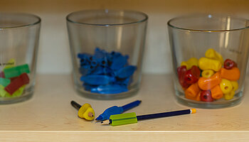 Drei Gläser mit farblich sortierten Griff- und Schreibhilfen stehen auf einem Regal; davor liegen Stifte und Aufsätze.