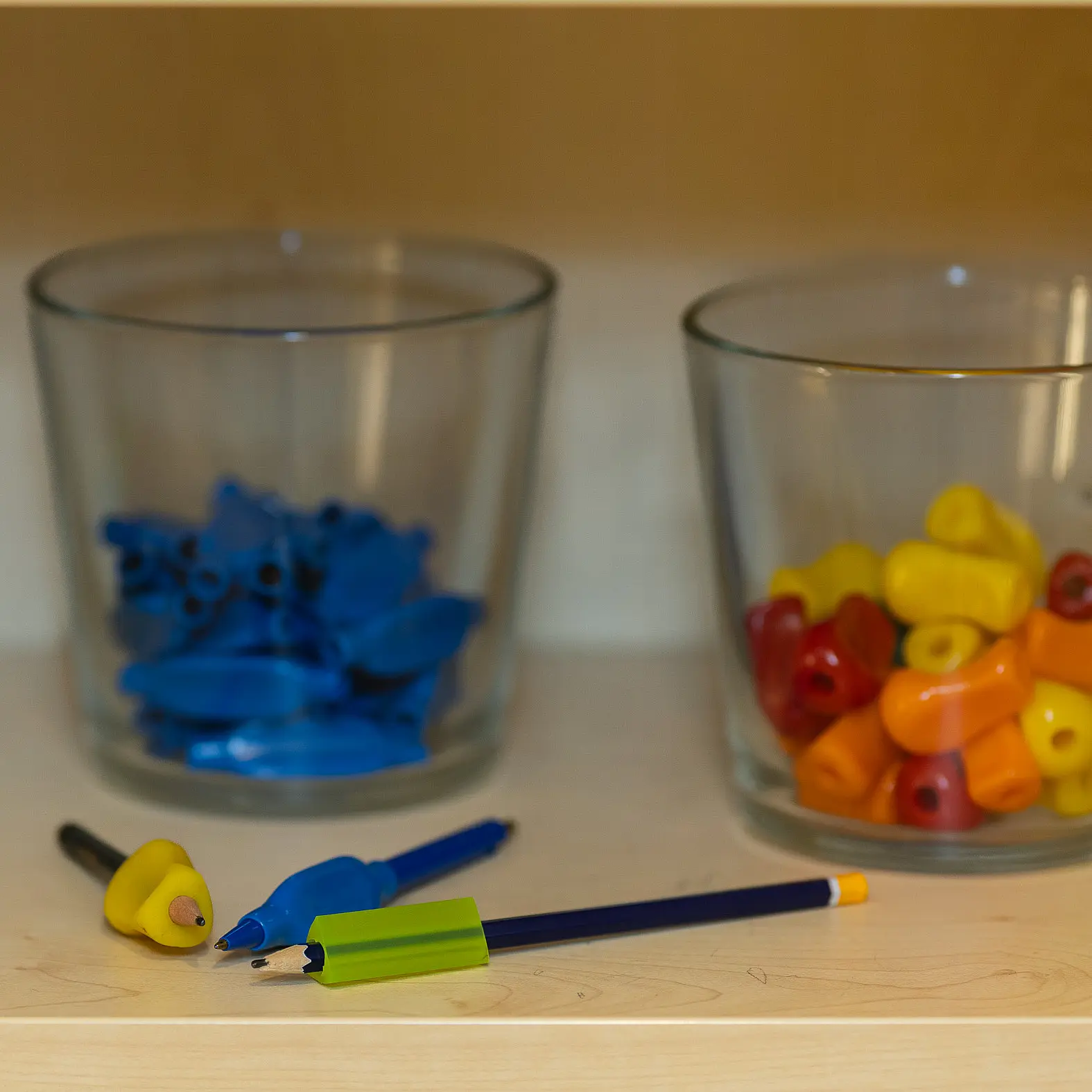 Drei Gläser mit farblich sortierten Griff- und Schreibhilfen stehen auf einem Regal; davor liegen Stifte und Aufsätze.