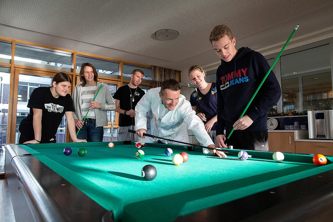 Jugendliche spielen gemeinsam Billard, begleitet von einem ärztlichen Mitarbeiter in weißem Kittel.