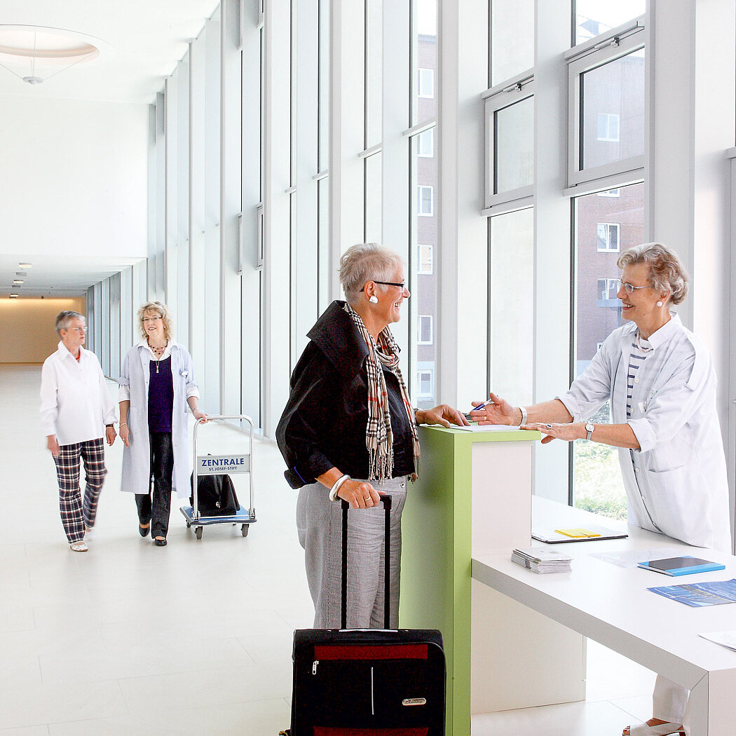 Empfangssituation an der Magistrale des St. Josef-Stifts: Eine Mitarbeiterin begrüßt eine Patientin mit Koffer am Counter, weitere Patient:innen und Mitarbeitende sind im Hintergrund zu sehen.