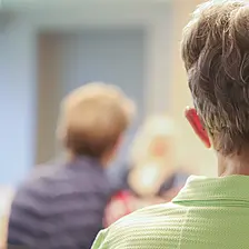 Teilnehmende sitzen in einem Raum und hören einem Angebot zu; im Vordergrund ist eine Person von hinten zu sehen, im Hintergrund sind weitere Personen unscharf erkennbar.