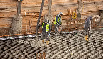 Drei Bauarbeitende verteilen frischen Beton auf einer dicht verlegten Stahlbewehrung. Ein Schlauch der Betonpumpe führt von oben auf die Fläche.