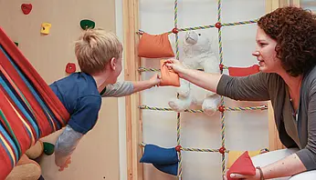 Ein Kind greift aus einer Hängematte nach einem kleinen Kissen, das ihm von einer Therapeutin gereicht wird; im Hintergrund sind Kletter- und Spielmaterialien zu sehen.