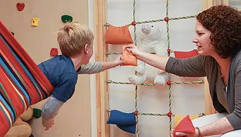 Ein Kind greift aus einer Hängematte nach einem kleinen Kissen, das ihm von einer Therapeutin gereicht wird; im Hintergrund sind Kletter- und Spielmaterialien zu sehen.