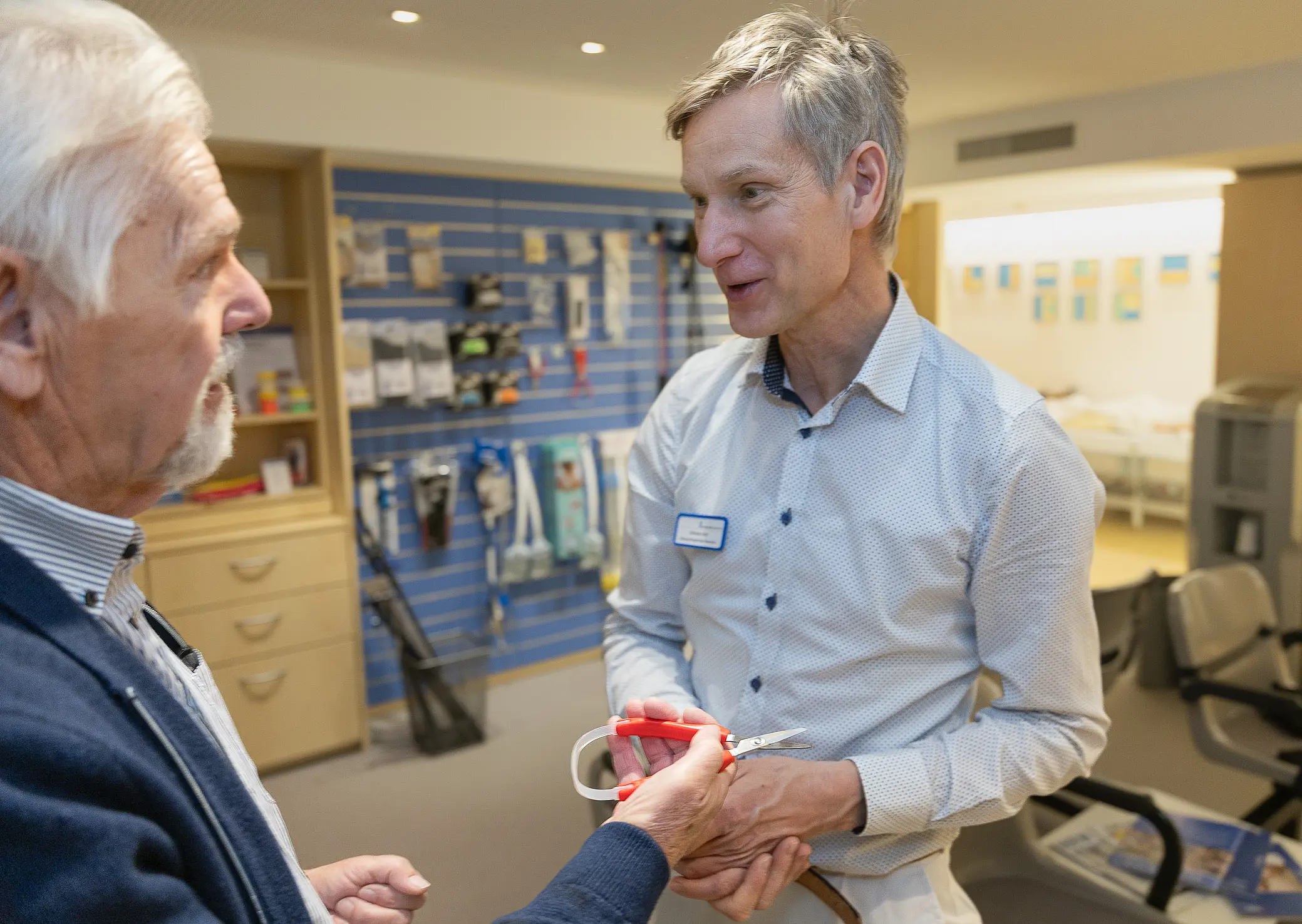 Fachkraft erläutert einem älteren Mann ein Hilfsmittel in einem Beratungsbereich mit Sitzgelegenheiten und Produktwand.