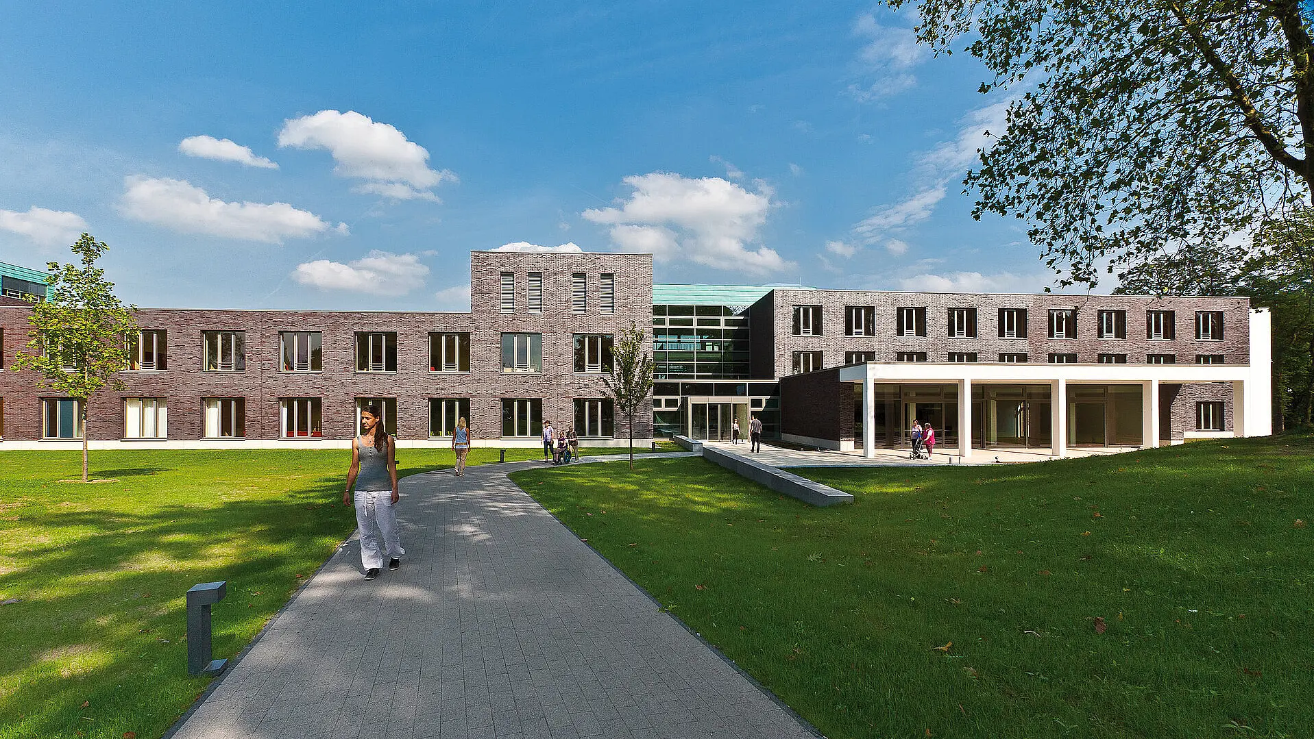 Außenansicht des Reha-Zentrums des St. Josef-Stifts mit begrüntem Gelände und Gehweg, auf dem Menschen unterwegs sind.