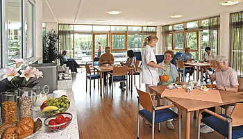 Heller Speiseraum mit gedeckten Tischen, sitzenden Patient:innen und Servicekraft, große Fenster mit Blick ins Grüne.