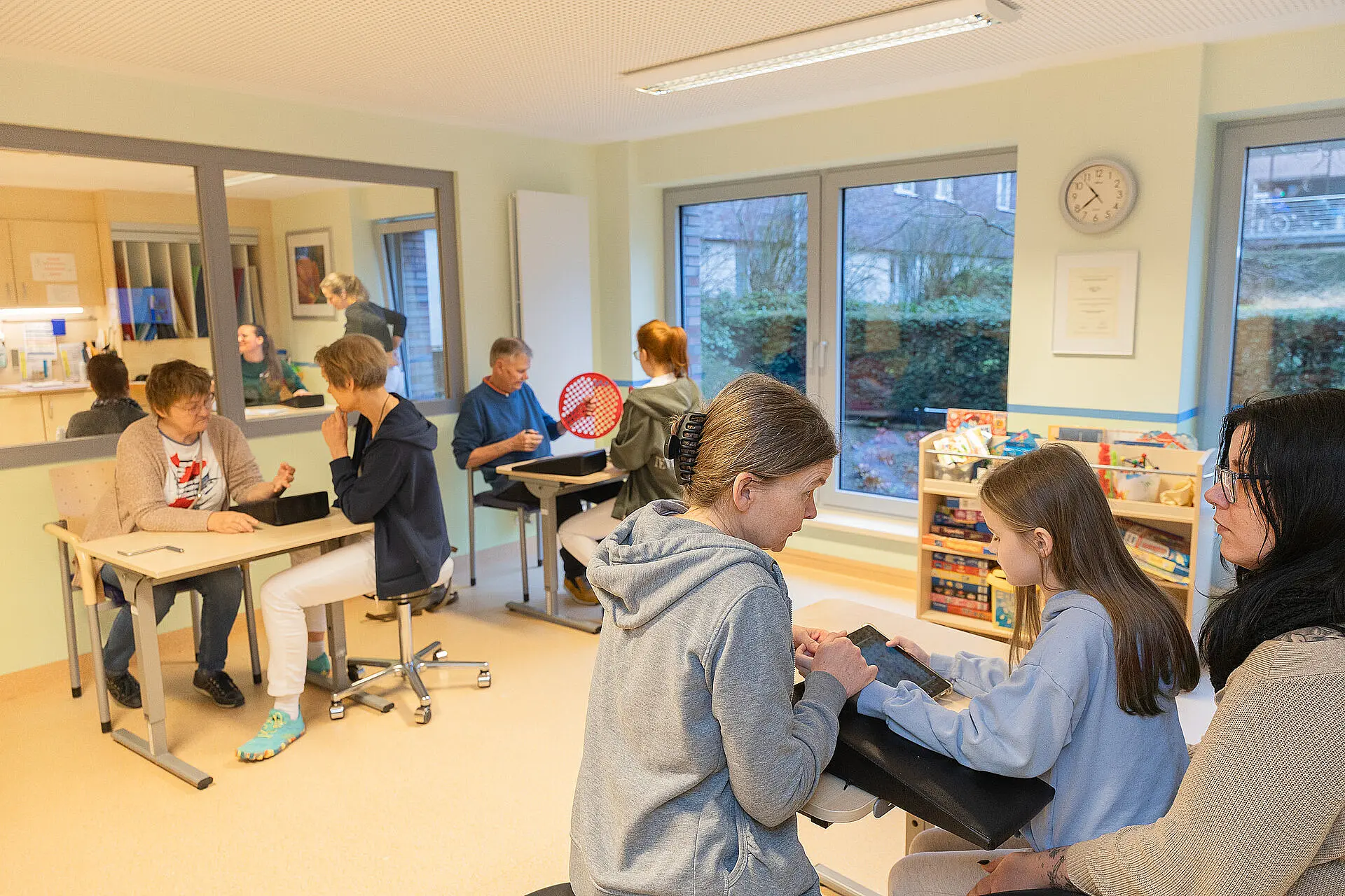 Großer Therapieraum mit mehreren Arbeitsplätzen: Eine Therapeutin begleitet eine erwachsene Patientin bei einer feinmotorischen Übung, während im selben Raum weitere Erwachsene und ein Kind an separaten Tischen therapeutische Aufgaben bearbeiten.