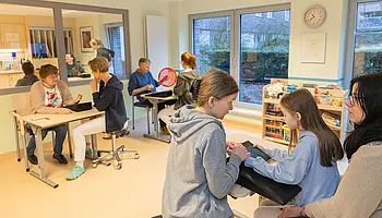 Mehrere Patient:innen unterschiedlichen Alters arbeiten in einem hellen Ergotherapieraum an Tischen mit therapeutischen Materialien; im Vordergrund unterstützt eine Therapeutin ein Kind bei einer Übung, im Hintergrund finden parallel weitere Therapiesituationen statt.