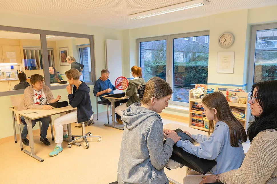 Großer Therapieraum mit mehreren Arbeitsplätzen: Eine Therapeutin begleitet eine erwachsene Patientin bei einer feinmotorischen Übung, während im selben Raum weitere Erwachsene und ein Kind an separaten Tischen therapeutische Aufgaben bearbeiten.