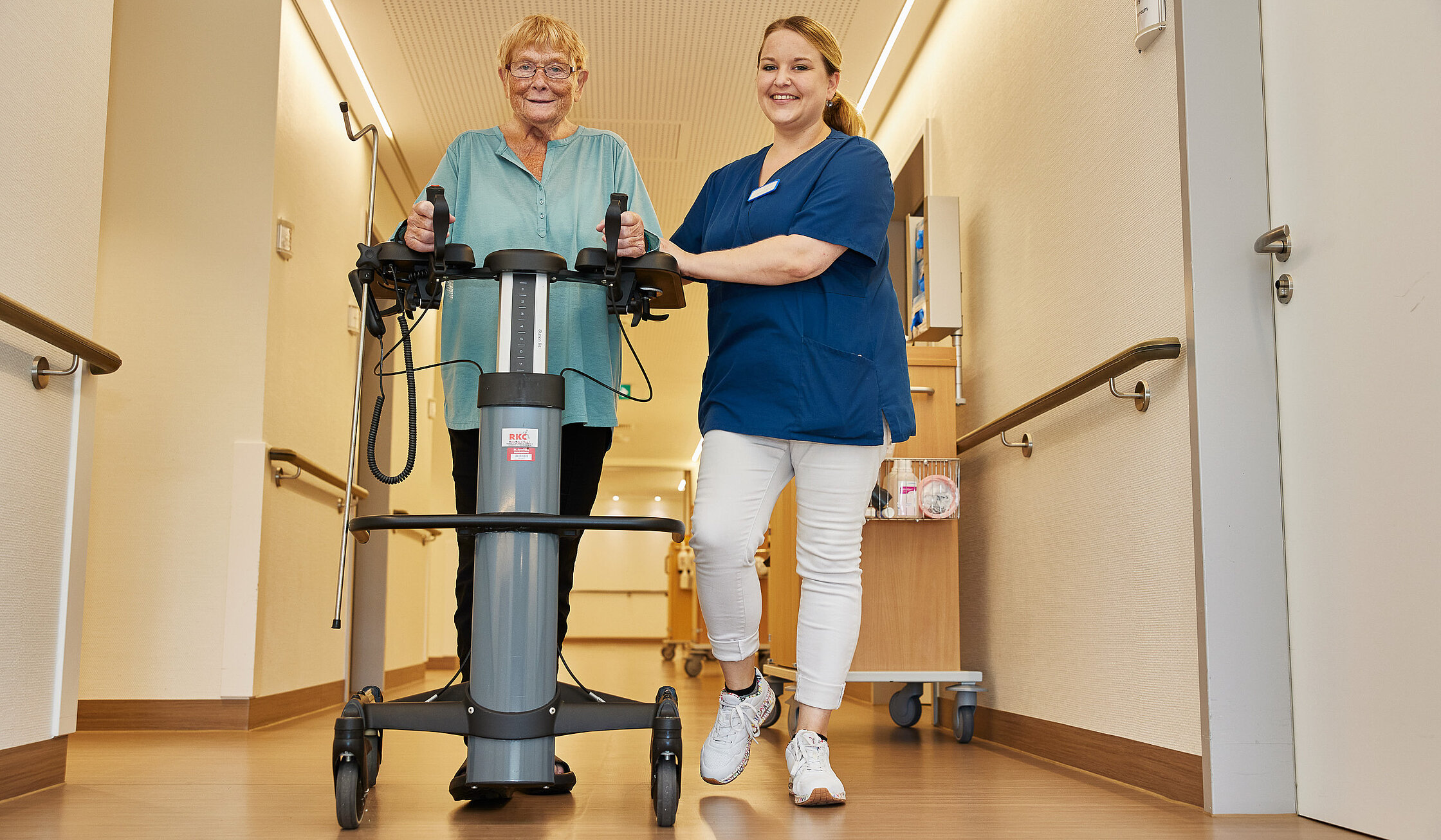 Eine Pflegefachkraft begleitet eine ältere Patientin beim Gehen mit einem Mobilisationsgerät auf dem Stationsflur.