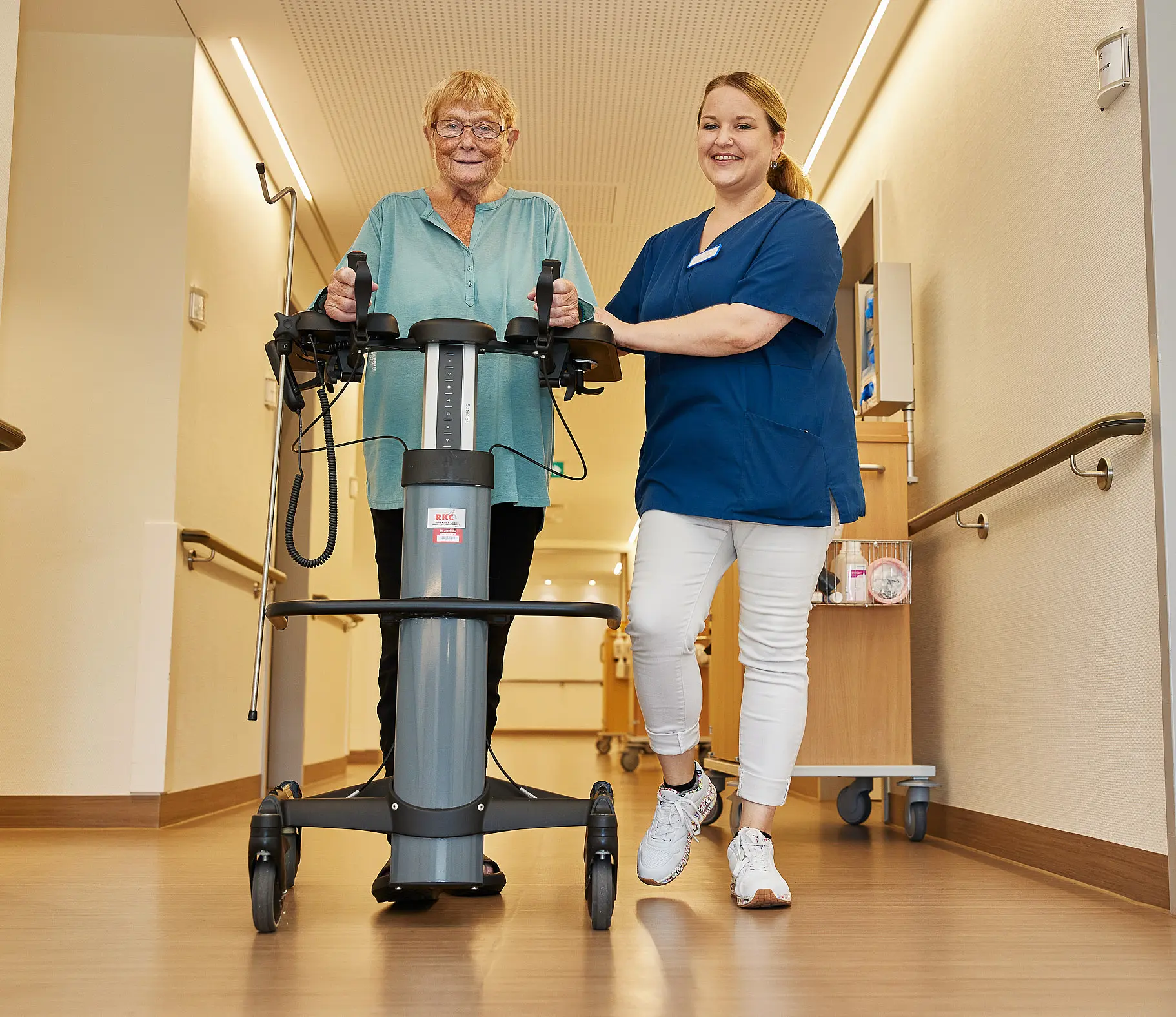 Eine Pflegefachkraft begleitet eine ältere Patientin beim Gehen mit einem Mobilisationsgerät auf dem Stationsflur.