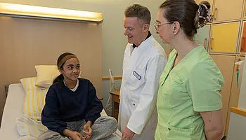 Ein Kind sitzt auf einem Krankenhausbett und spricht mit Dr. PD Daniel Windschall. Eine Mitarbeiterin steht neben ihm und hört zu.