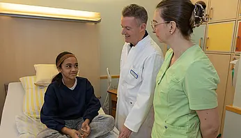 Ein Kind sitzt auf einem Krankenhausbett und spricht mit Dr. PD Daniel Windschall. Eine Mitarbeiterin steht neben ihm und hört zu.