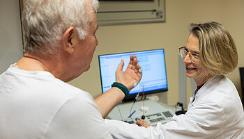 Ärztin spricht mit einem Patienten und erklärt Untersuchungsergebnisse an einem Computerarbeitsplatz.