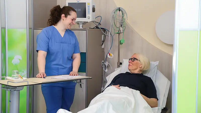 Eine Patientin liegt im Aufwachraum und wird von einer Pflegefachkraft freundlich angesprochen. Medizinische Geräte sind im Hintergrund zu sehen.
