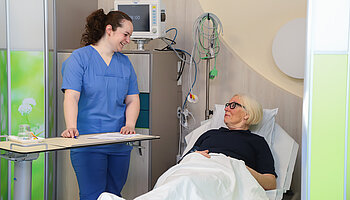 Eine Patientin liegt im Aufwachraum und wird von einer Pflegefachkraft freundlich angesprochen. Medizinische Geräte sind im Hintergrund zu sehen.