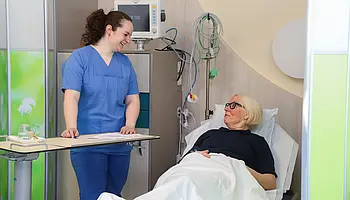 Eine Patientin liegt im Aufwachraum und wird von einer Pflegefachkraft freundlich angesprochen. Medizinische Geräte sind im Hintergrund zu sehen.