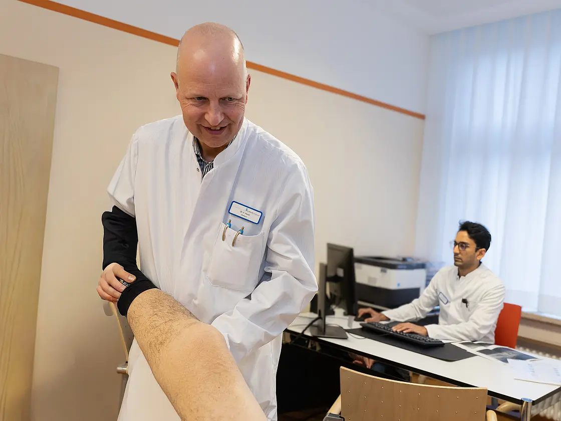 Ein Arzt in weißem Kittel untersucht das Bein eines Patienten. Im Hintergrund sitzt ein weiterer Arzt an einem Schreibtisch und arbeitet am Computer.