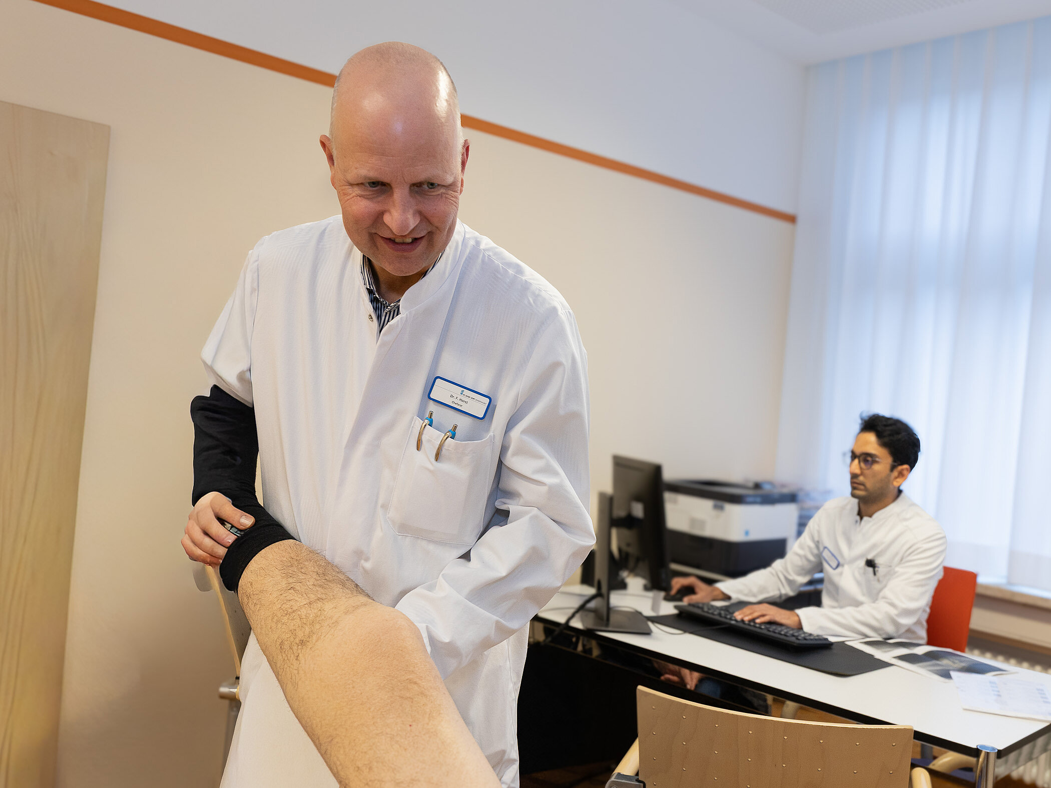 Ein Arzt in weißem Kittel untersucht das Bein eines Patienten. Im Hintergrund sitzt ein weiterer Arzt an einem Schreibtisch und arbeitet am Computer.