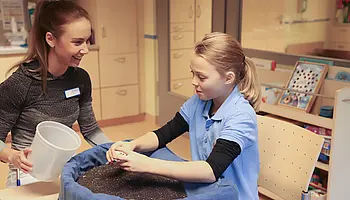 Ergotherapeutische Fachkraft begleitet ein Mädchen mit Kinderrheuma bei einer beidhändigen Übung in einer mit Rapssamen gefüllten Schale an einem Tisch.