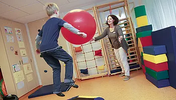 Ein Kind springt auf eine weiche Matte und spielt mit einem großen roten Ball, während eine Therapeutin gegenübersteht.