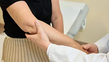 Nahaufnahme eines Ellenbogens, der von einer Hand im weißen Arztkittel vorsichtig abgetastet wird.