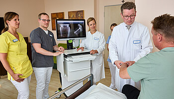 Dr. med. Ansgar Platte steht mit mehreren Mitarbeitenden an einem Patientenbett; ein mobiler Monitor mit Röntgenaufnahmen ist neben dem Bett positioniert.