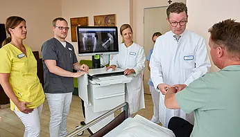 Dr. med. Ansgar Platte steht mit mehreren Mitarbeitenden an einem Patientenbett; ein mobiler Monitor mit Röntgenaufnahmen ist neben dem Bett positioniert.