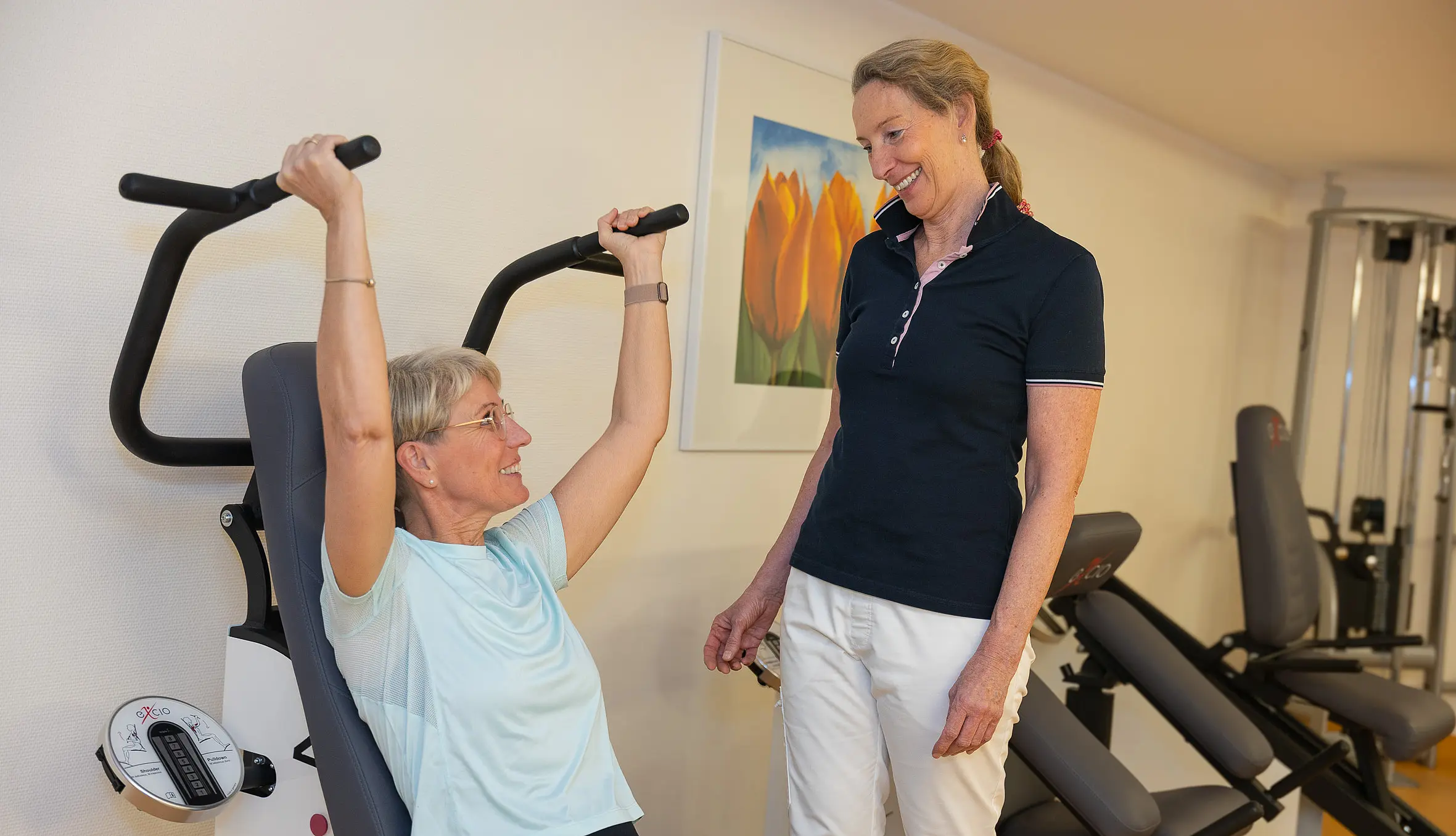Eine Patientin trainiert sitzend an einem Gerät mit Überkopfgriffen, eine Therapeutin beobachtet die Übung und gibt Hinweise.