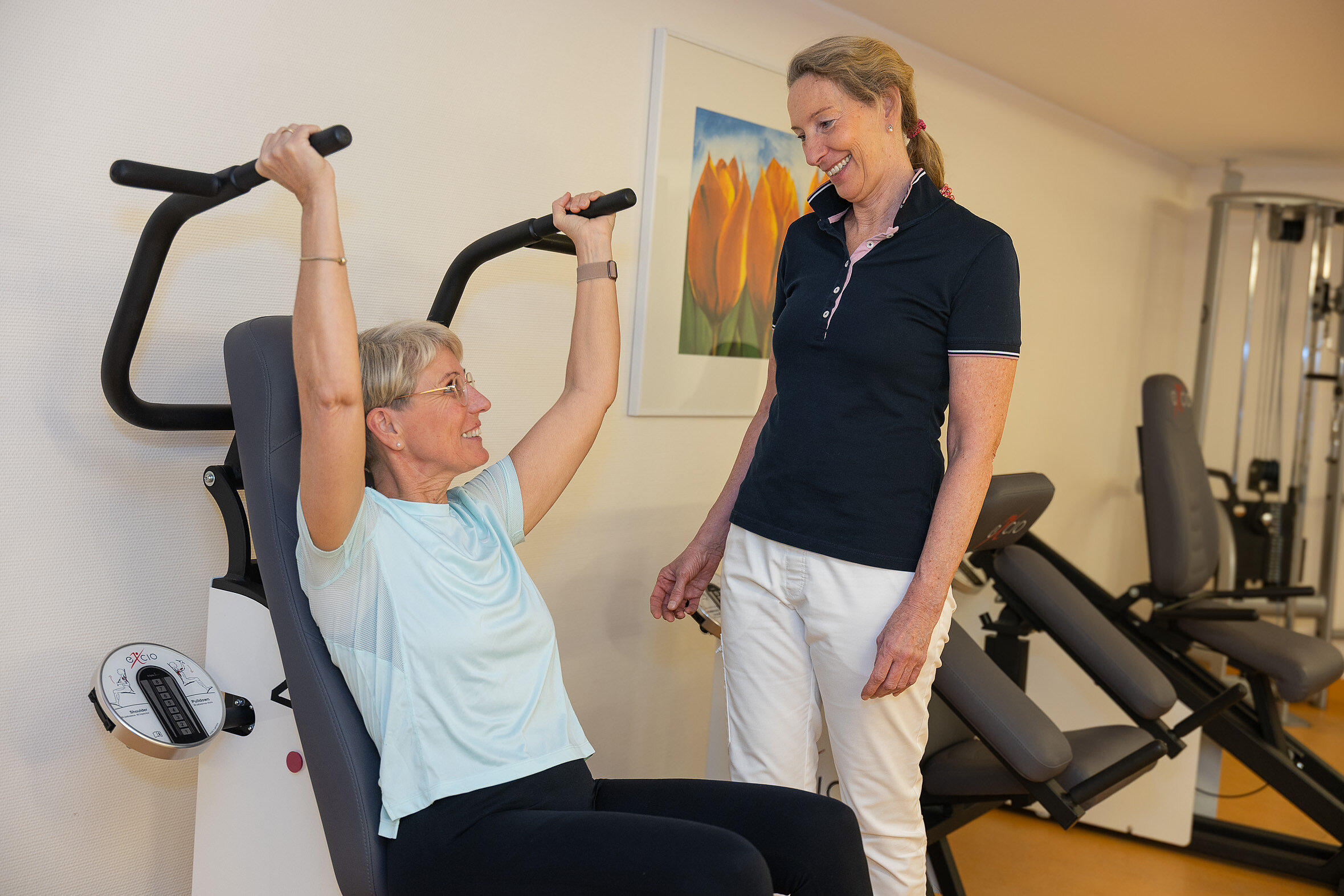 Eine Patientin trainiert sitzend an einem Gerät mit Überkopfgriffen, eine Therapeutin beobachtet die Übung und gibt Hinweise.