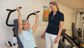 Eine Patientin trainiert sitzend an einem Gerät mit Überkopfgriffen, eine Therapeutin beobachtet die Übung und gibt Hinweise.
